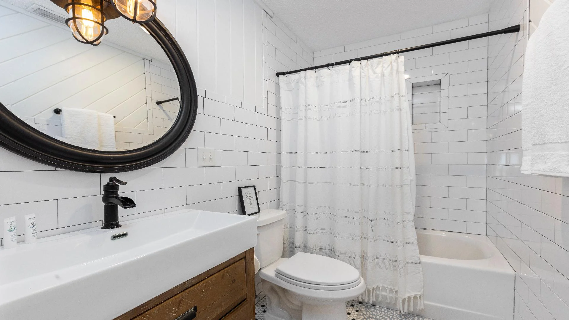Bathroom with white subway tiles, a black oval mirror, wood vanity, and a bathtub behind a fringed shower curtain.