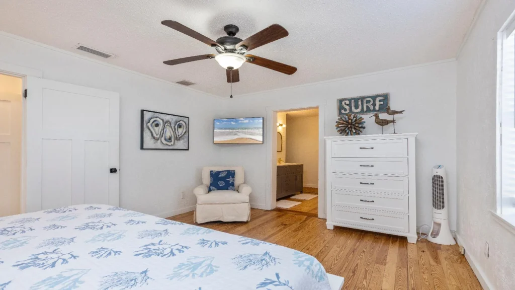Bedroom with hardwood floors, white dresser topped with coastal decor, and a ceiling fan overhead.