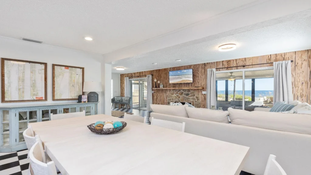 Light wood dining table seats eight on black-and-white checkered floor beside living room with stone fireplace and ocean view.