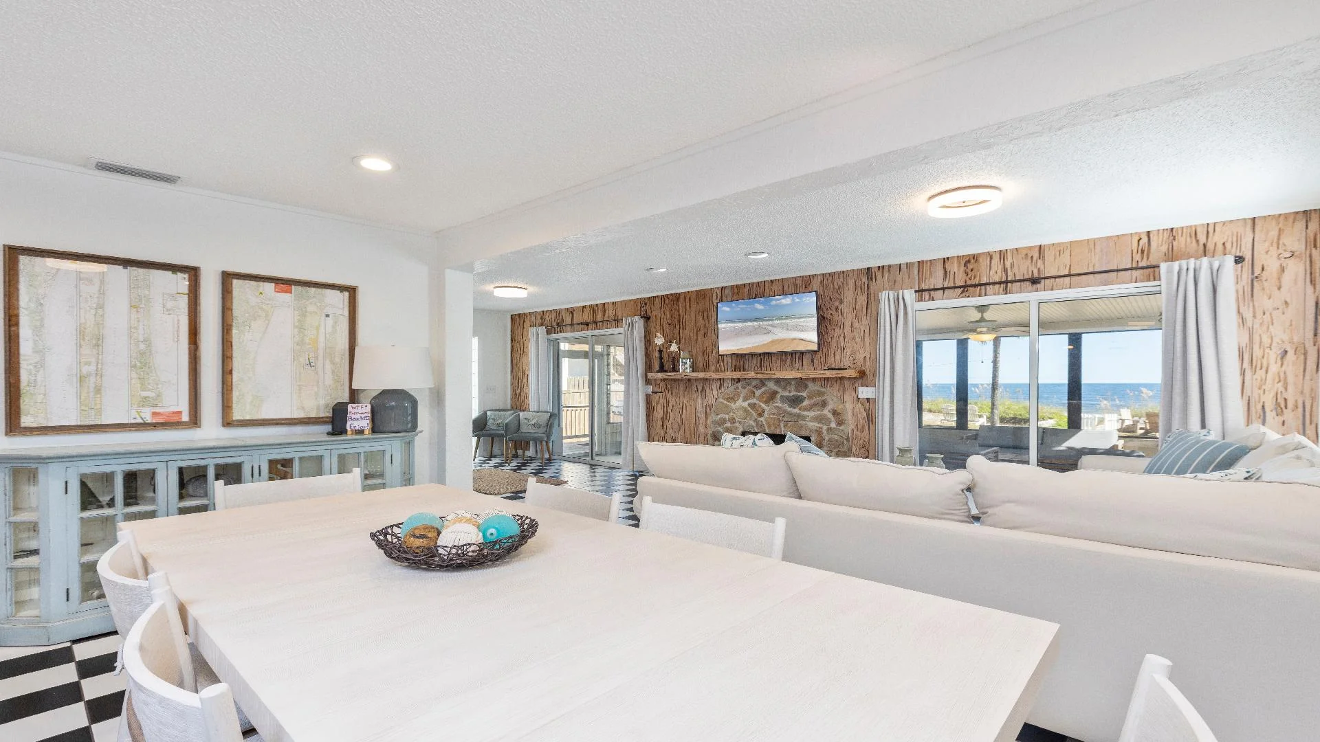 Light wood dining table seats eight on black-and-white checkered floor beside living room with stone fireplace and ocean view.
