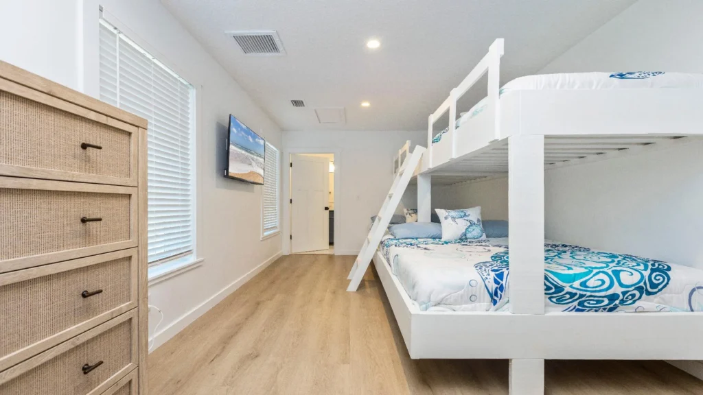 Bunk bedroom with white wooden frame, blue and white patterned bedding, wall-mounted TV, and light wood flooring.