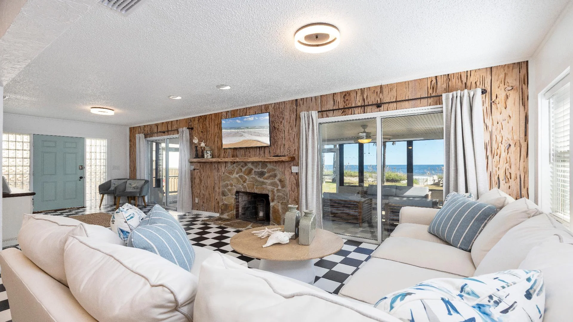 Living room with black-and-white checkered floor, stone fireplace, wood-paneled wall, and ocean views through sliding glass doors.