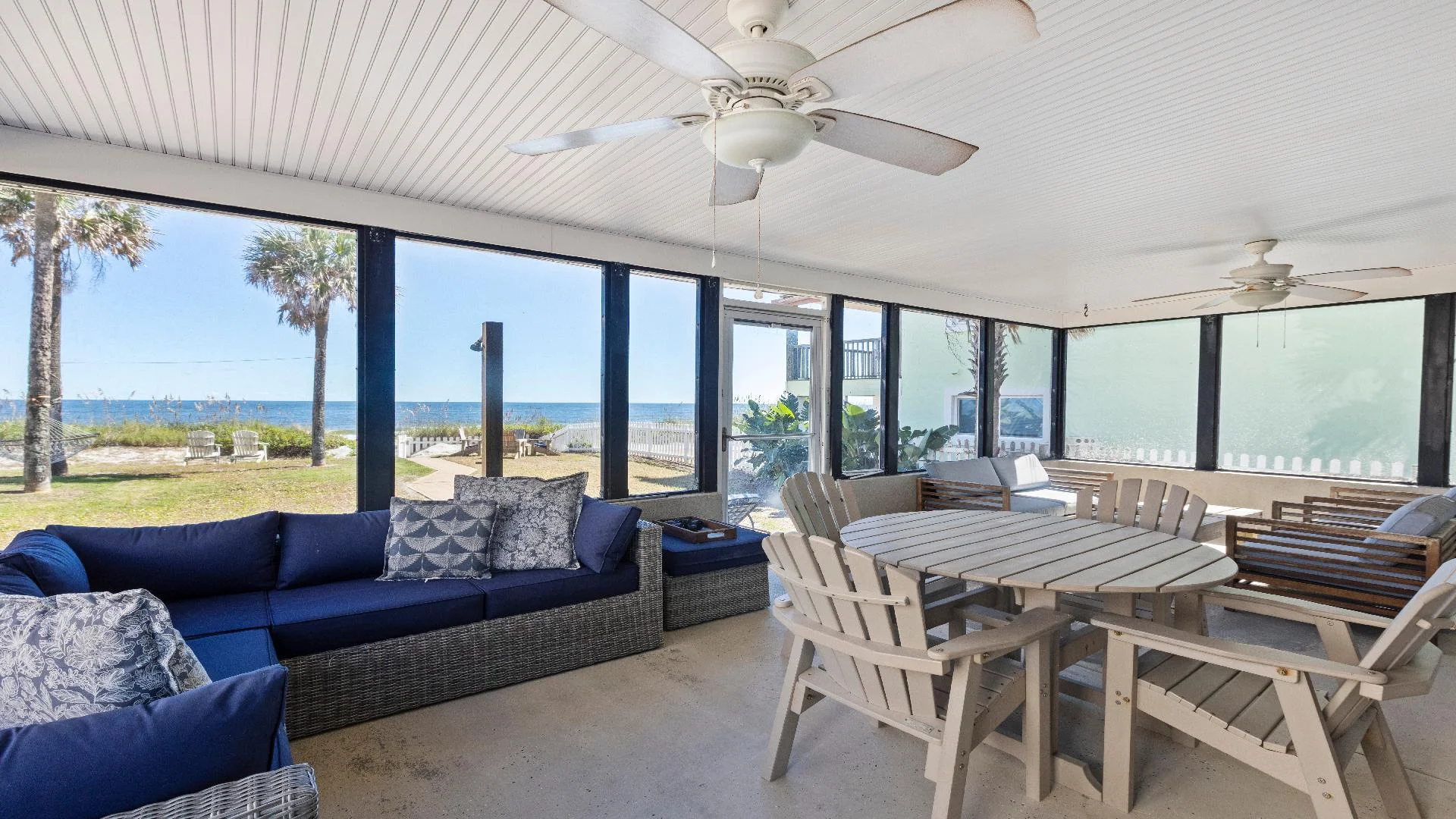 Screened porch with navy wicker sectional, beige Adirondack chairs, ceiling fans, and ocean views through tall windows.