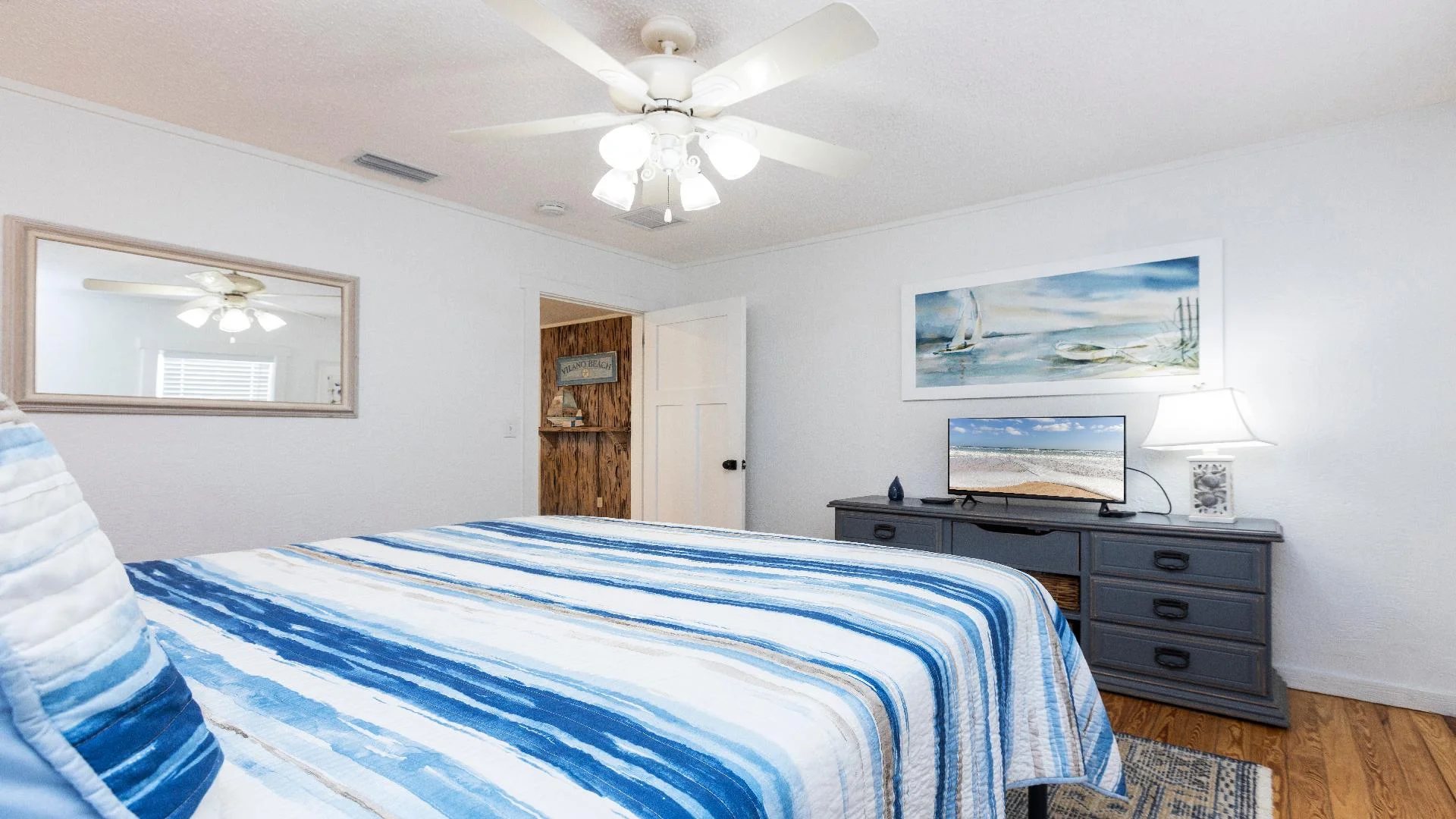 Bedroom with striped blue bedding, hardwood floors, dark gray dresser, and coastal painting above flat-screen TV.