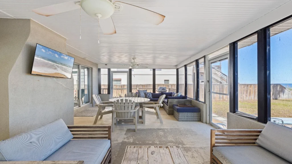 Screened porch with wooden Adirondack chairs around a round table, wicker sectional, ceiling fans, and ocean views.