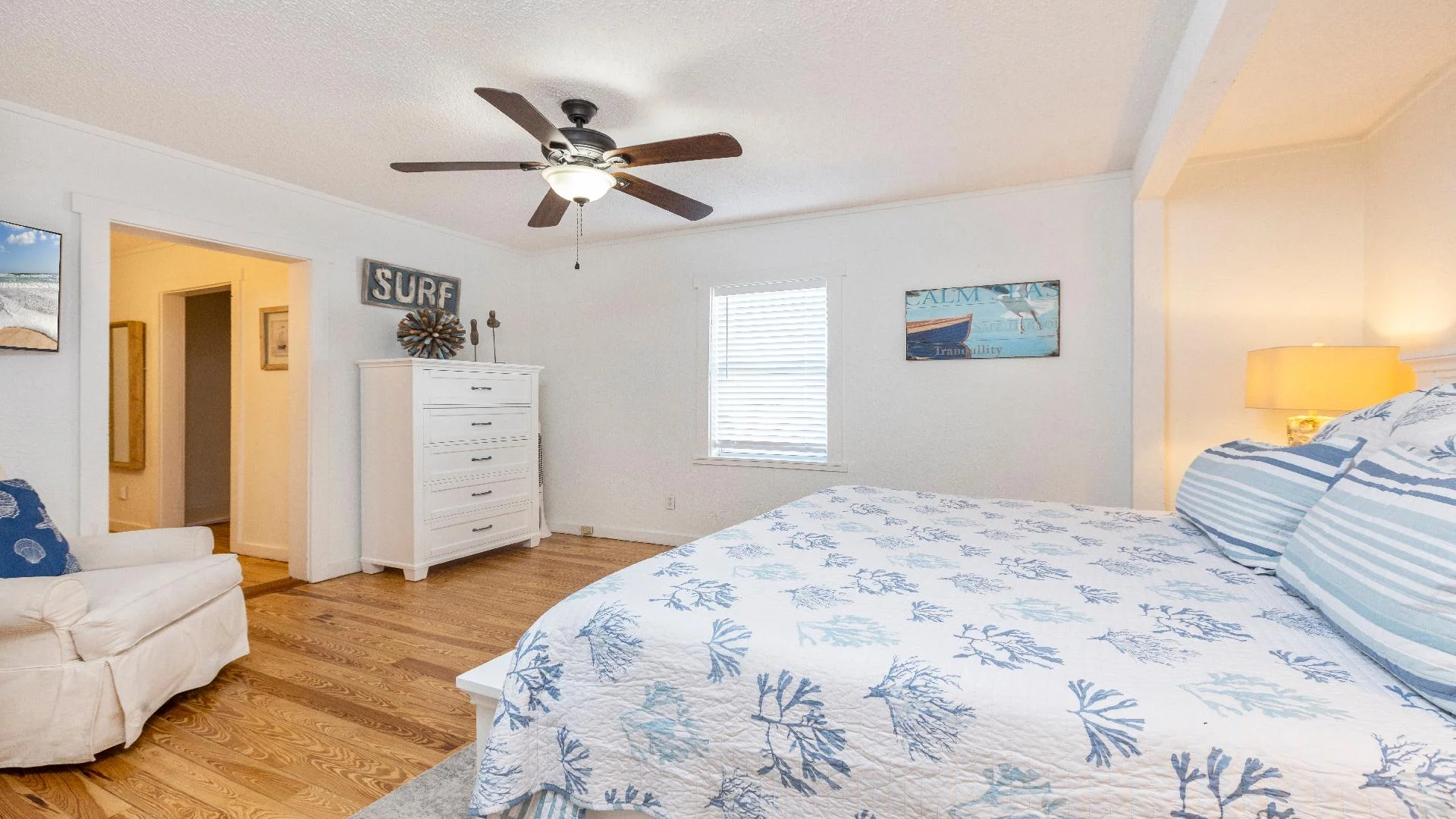 Bedroom with hardwood floors, white dresser, ceiling fan, and coastal-themed bedding and artwork.