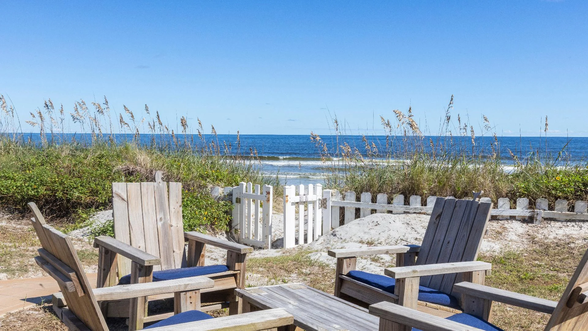 Weathered wooden Adirondack chairs with blue cushions encircle a low table near a white picket gate opening to the sandy beach.