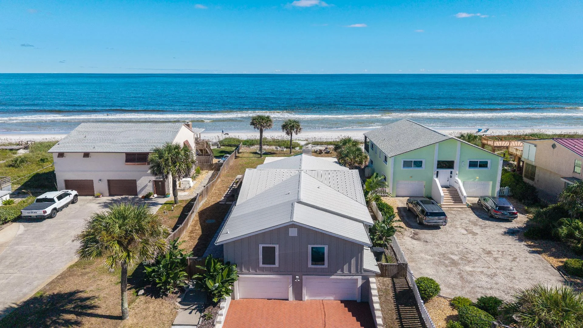 Oceanfront exterior view of the house with a metal roof, palm trees, and sandy beach shoreline beyond.