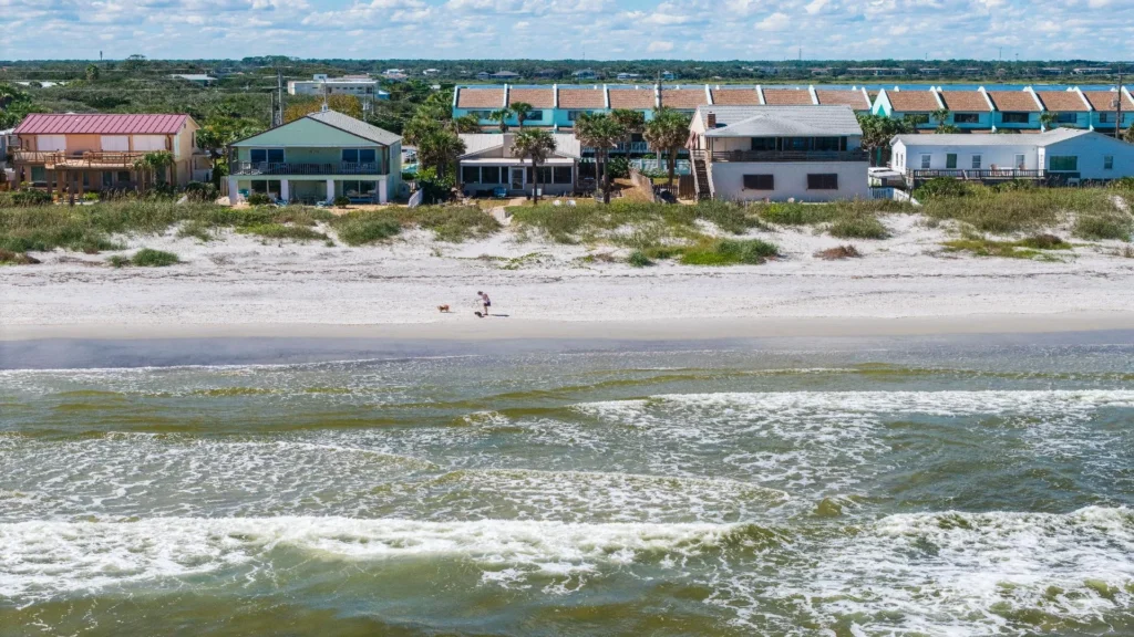 Oceanfront view of sandy beach with gentle waves, beachfront houses, and a person walking a dog.
