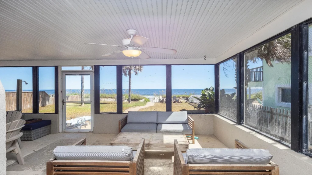 Screened porch with wood slat seating, striped cushions, a white ceiling fan, and ocean views through tall windows.