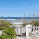 Open white picket gate leads through sand dunes with sea oats to the ocean shore under clear blue sky.