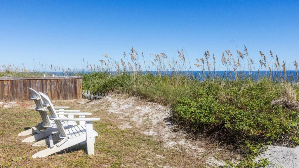 Two white Adirondack chairs face the ocean beyond sand dunes and tall grasses under a clear blue sky.