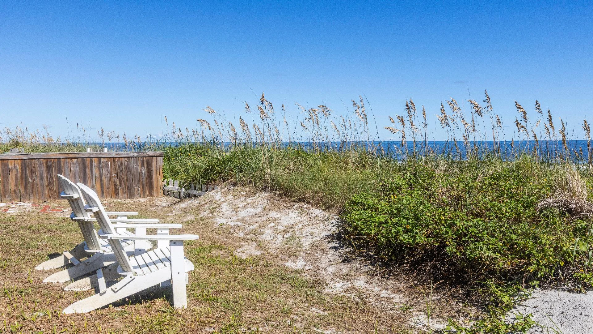Two white Adirondack chairs face the ocean beyond sand dunes and tall grasses under a clear blue sky.