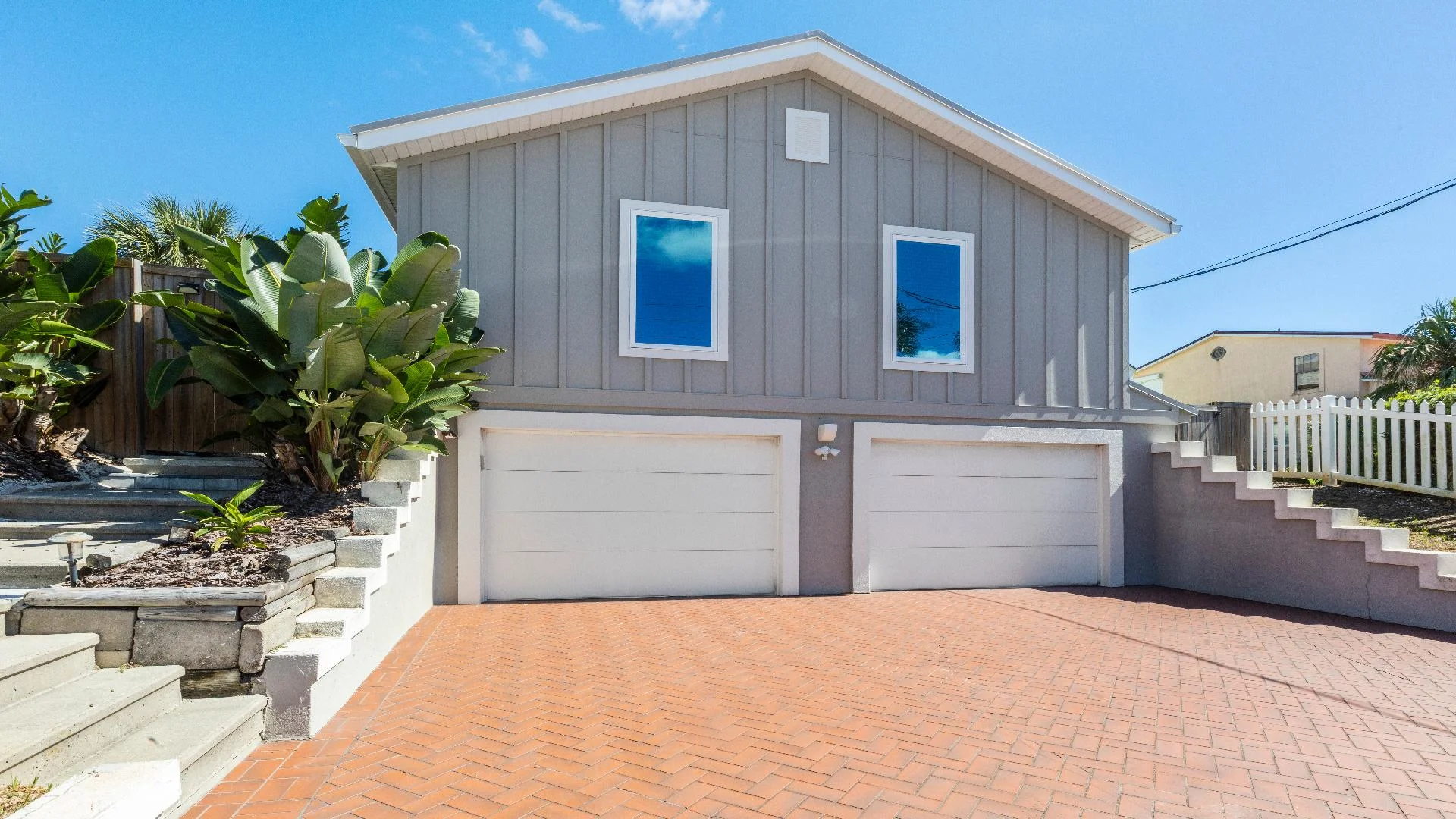 Exterior view of gray board-and-batten siding with two white garage doors and terracotta brick driveway.