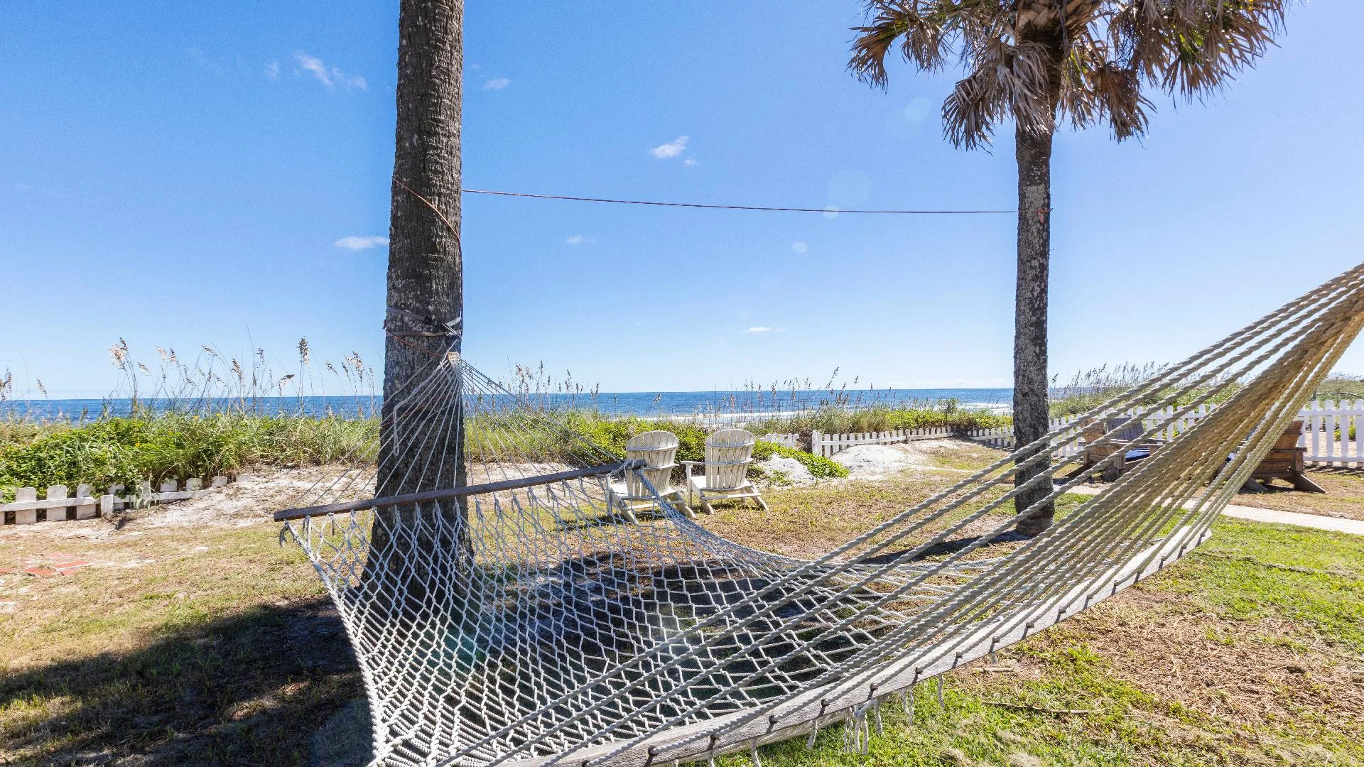 Rope hammock strung between palm trees overlooks sandy dunes and ocean under clear blue sky.