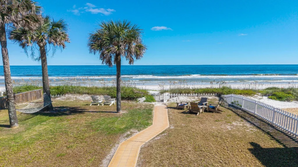 Oceanfront yard with a paved walkway, palm trees, hammock, Adirondack chairs, and a white picket fence gate.