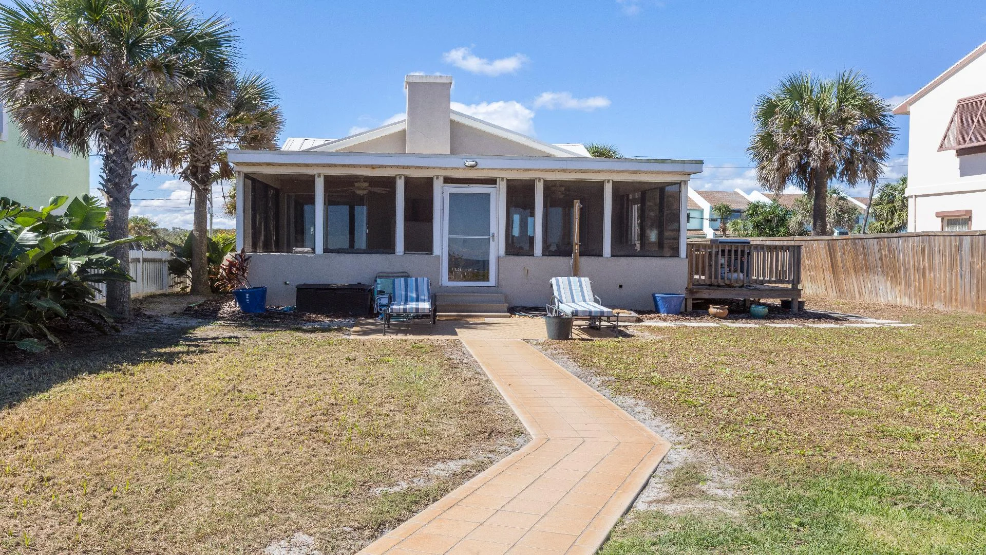 Backyard with tiled walkway leading to a screened porch, flanked by two striped lounge chairs and palm trees.
