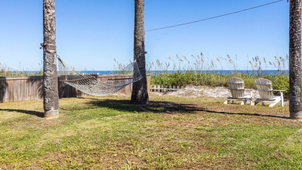 Oceanfront yard with a white rope hammock suspended between palm trees and two Adirondack chairs facing the water.
